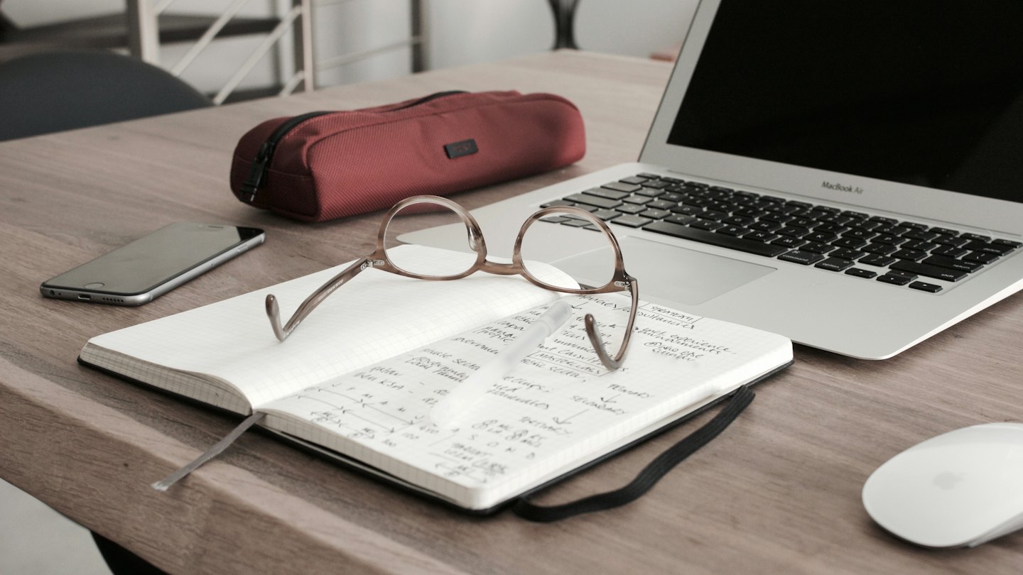 Laptop, notatnik, okulary i telefon na drewnianym biurku.
